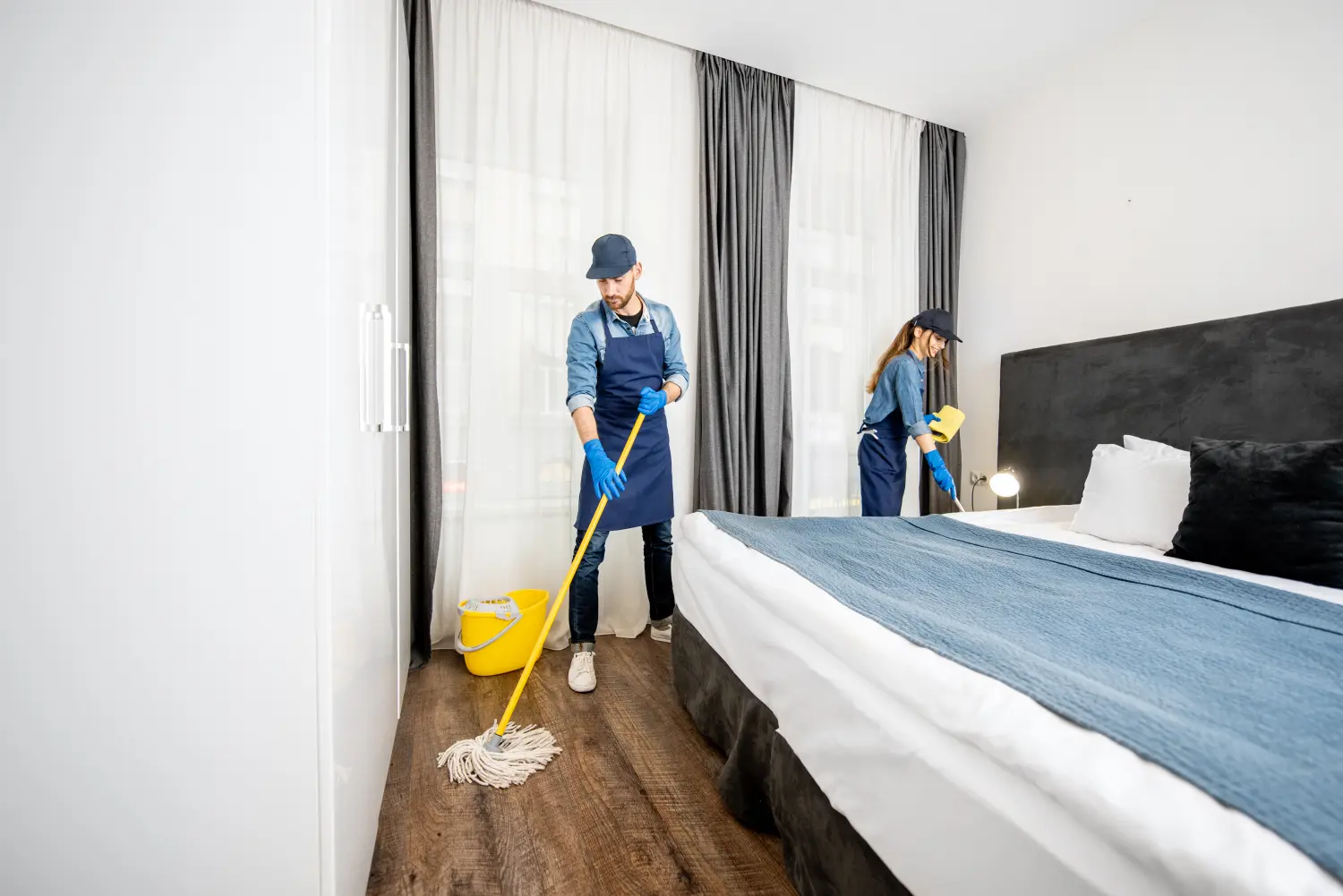 Professionelle Reinigungskräfte in Uniform wischen den Boden und wischen Staub von den Möbeln im Hotelzimmer